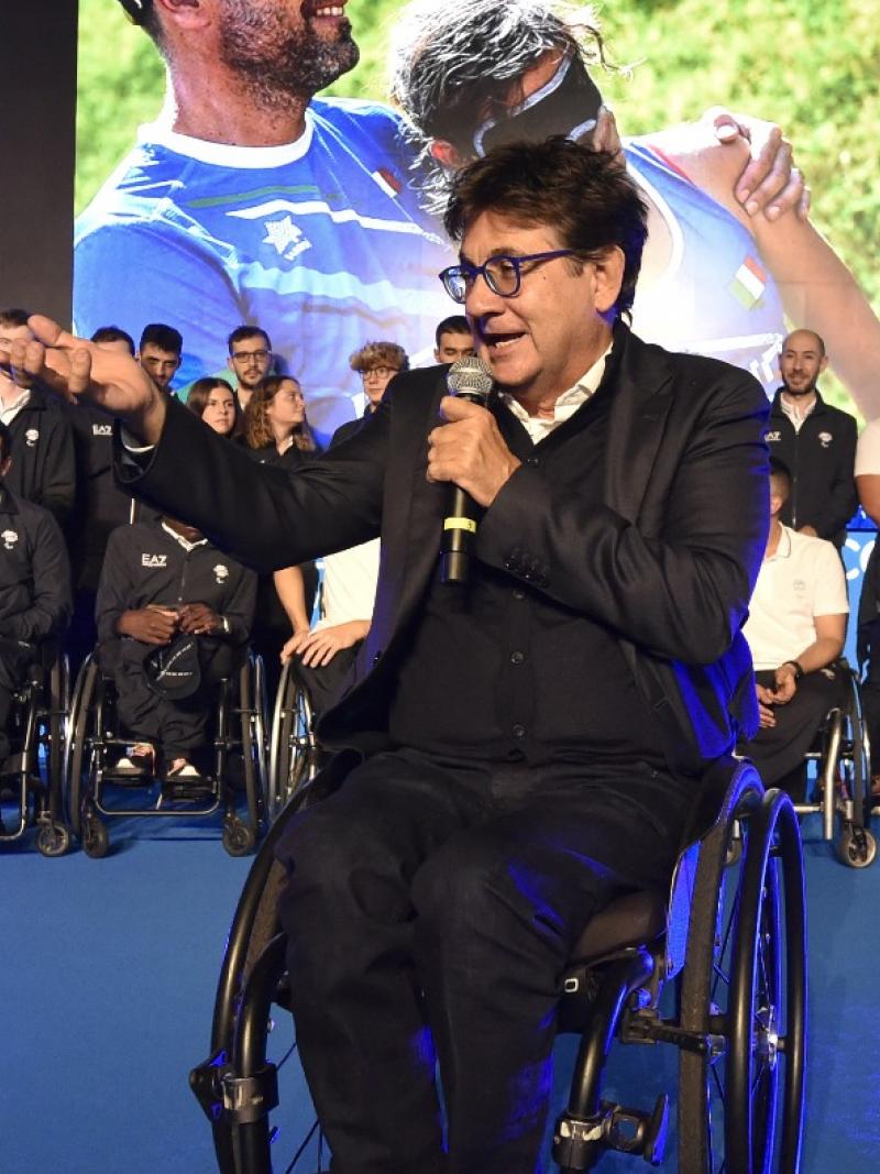 A man in a wheelchair speaks in a microphone on stage with a crowd of athletes behind him.